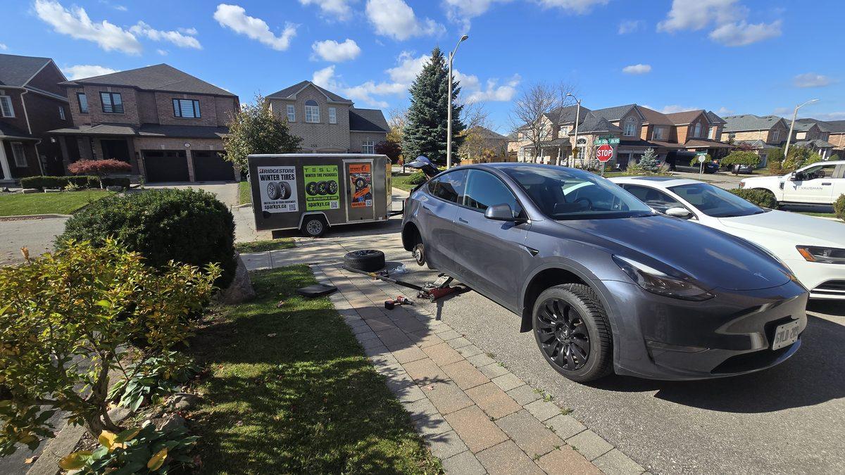 Grey Tesla Model Y on a jack mid winter tire change in a customer's driveway — rear wheel off, tools laid out, the Sparky X trailer with Bridgestone and Tesla winter package signage behind it