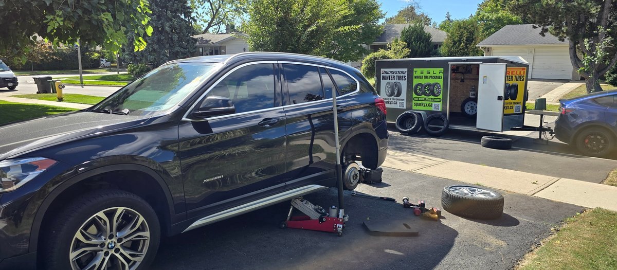 Sparky X mobile tire service at work — a black BMW X1 on a jack in a suburban driveway, rear wheel off and tools laid out, the branded Sparky X service trailer open behind it under a bright blue sky